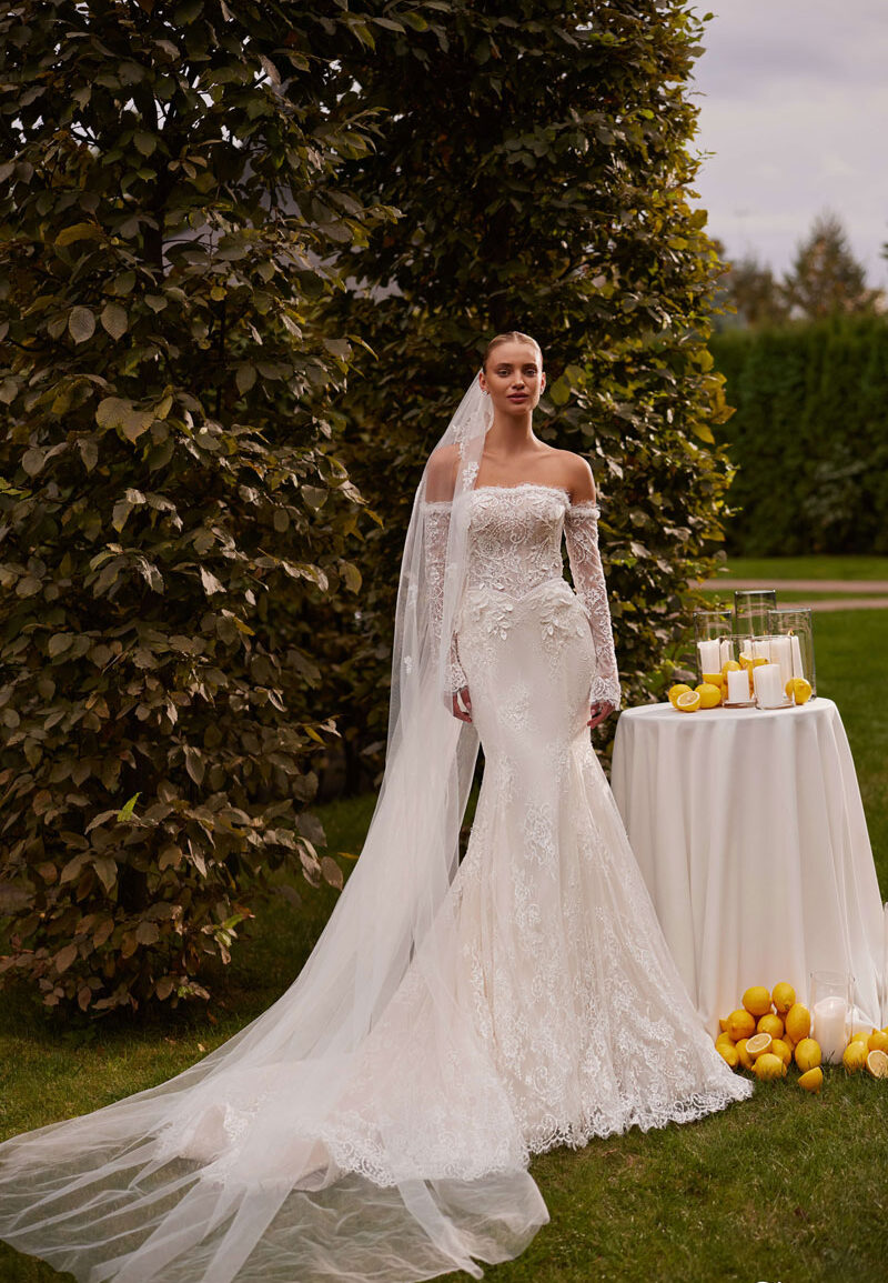 Bride wearing an off-the-shoulder lace mermaid wedding dress with a long train and veil, standing beside a candle-lit table at an outdoor garden venue near Liverpool, from The White Gallery Ramsbottom.