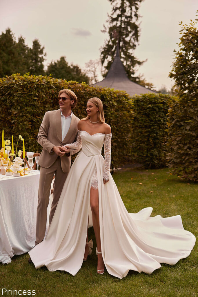 Bride wearing an elegant off-the-shoulder princess wedding dress with lace sleeves and thigh split, walking hand in hand with her partner at an outdoor celebration near Liverpool, from The White Gallery Ramsbottom.