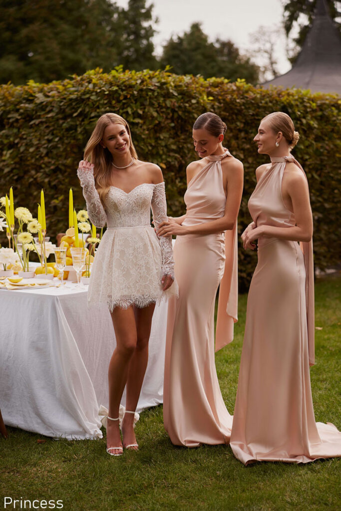 Bride wearing a short lace off-the-shoulder wedding dress with long sleeves, standing with bridesmaids in blush satin gowns at an outdoor reception near Liverpool, from The White Gallery Ramsbottom.